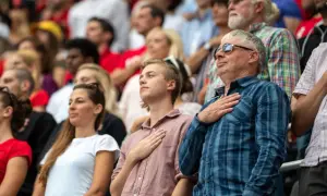 Widownia na stadionie w geście szacunku, z dłonią na sercu. Widać dumę i poczucie wspólnoty, prawdziwą **gloria** chwili.