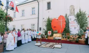 Uroczysta procesja Bożego Ciała przed kościołem. Wierni w białych szatach, wielkie serce z krzyżem i transparent "Król Miłości i Pokoju".