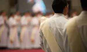 Diakon w białej szacie z żółtą stułą, przygotowuje się do uroczystości. W tle widać innych duchownych.