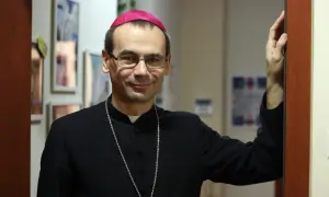 Uśmiechnięty, najmłodszy biskup w Polsce, w czarnej sutannie i z różowym zucchetto na głowie, opiera się o framugę drzwi.