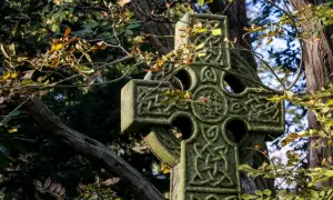 Kamienny krzyż celtycki z misternymi wzorami, otoczony jesiennymi liśćmi i gałęziami drzew.
