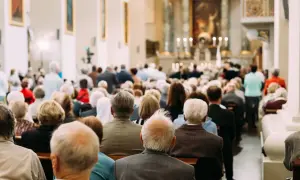Ludzie siedzą w kościele podczas mszy wielkosobotniej. Widać ołtarz z zapalonymi świecami i obrazy na ścianach.