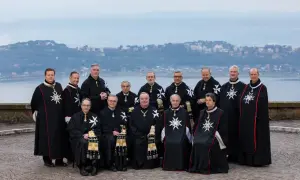 Uroczyste zgromadzenie członków zakonu maltańskiego w czarnych płaszczach z białymi krzyżami, na tle jeziora i miasta.