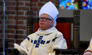 Biskup w białej mitrze i ozdobnej szacie z krzyżem, siedzi na drewnianym tronie.
