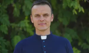 Młody ksiądz z osiedla w granatowej koszuli z kołnierzykiem, uśmiecha się na tle zieleni.
