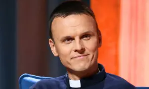 Uśmiechnięty ksiądz z osiedla, młody duchowny w sutannie, z uwagą słucha rozmówcy.