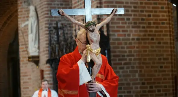 Ksiądz w czerwonej szacie adoruje krzyż z figurą Chrystusa. Trwa liturgia Wielkiego Piątku, która jest wyrazem głębokiej refleksji.