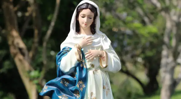 Figurka Matki Boskiej w błękitnym płaszczu ze złotymi zdobieniami. W tle drzewa. Idealna na 3 maja święto kościelne.