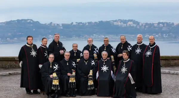 Uroczyste zgromadzenie członków zakonu maltańskiego w czarnych płaszczach z białymi krzyżami, na tle jeziora i miasta.