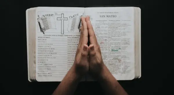 Dłonie złożone w modlitwie nad otwartą Biblią, z widocznymi rozdziałami o Starym i Nowym Testamencie, symbolizują głębokie rozważania i psalmy uwielbienia.