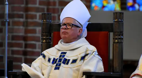 Biskup w białej mitrze i ozdobnej szacie z krzyżem, siedzi na drewnianym tronie.