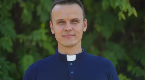 Młody ksiądz z osiedla w granatowej koszuli z kołnierzykiem, uśmiecha się na tle zieleni.