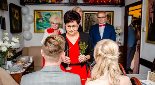 Krótkie błogosławieństwo pary młodej przez starszą panią z gałązką. W tle goście i dekoracje.