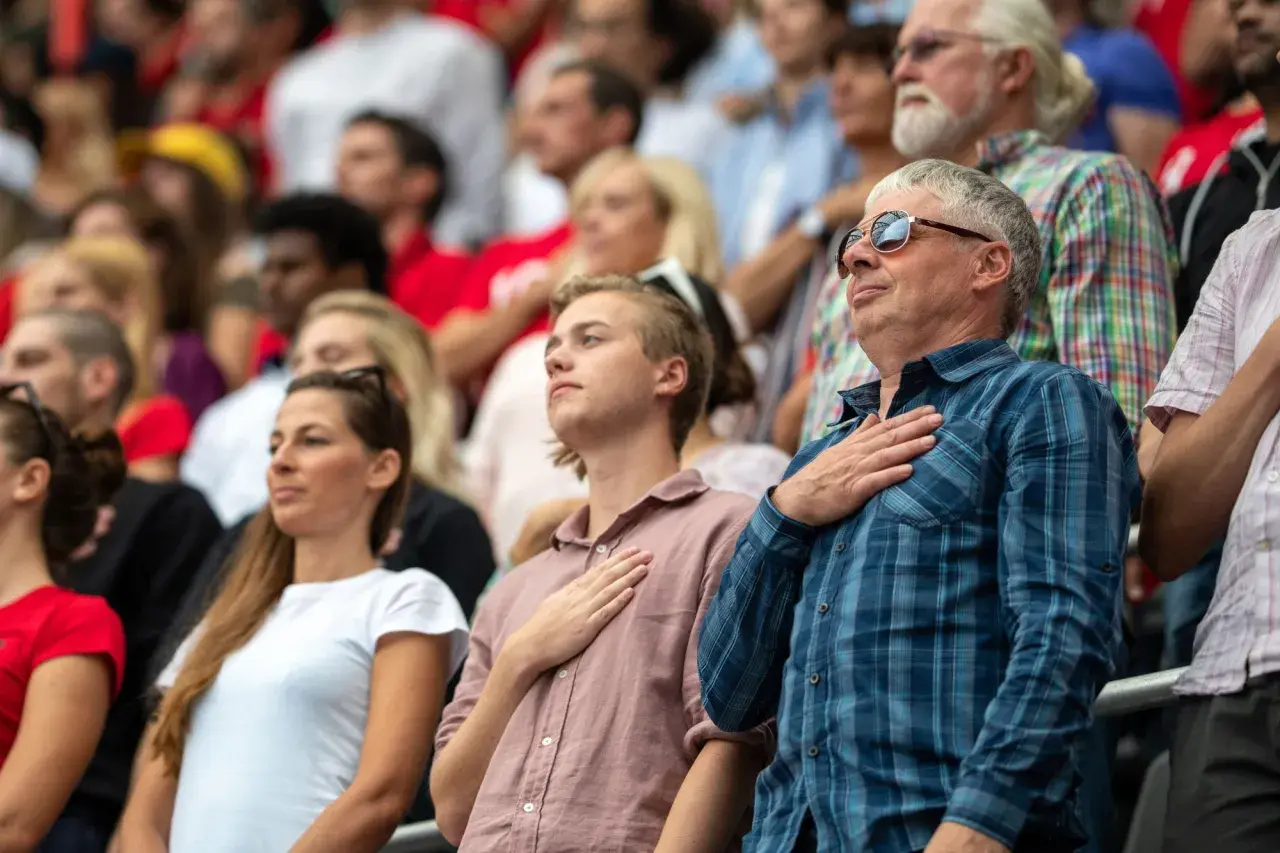 Widownia na stadionie w geście szacunku, z dłonią na sercu. Widać dumę i poczucie wspólnoty, prawdziwą **gloria** chwili.