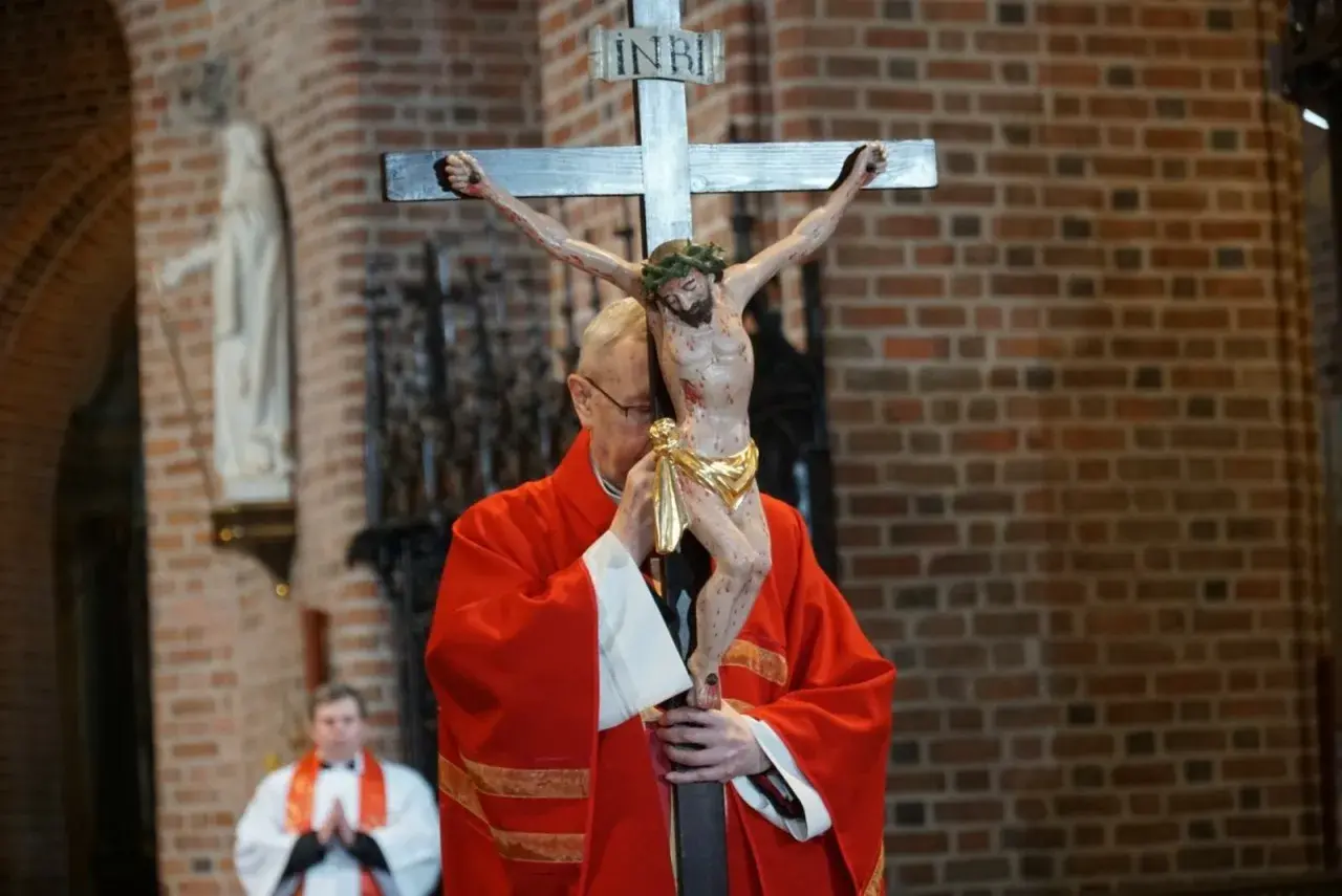 Ksiądz w czerwonej szacie adoruje krzyż z figurą Chrystusa. Trwa liturgia Wielkiego Piątku, która jest wyrazem głębokiej refleksji.