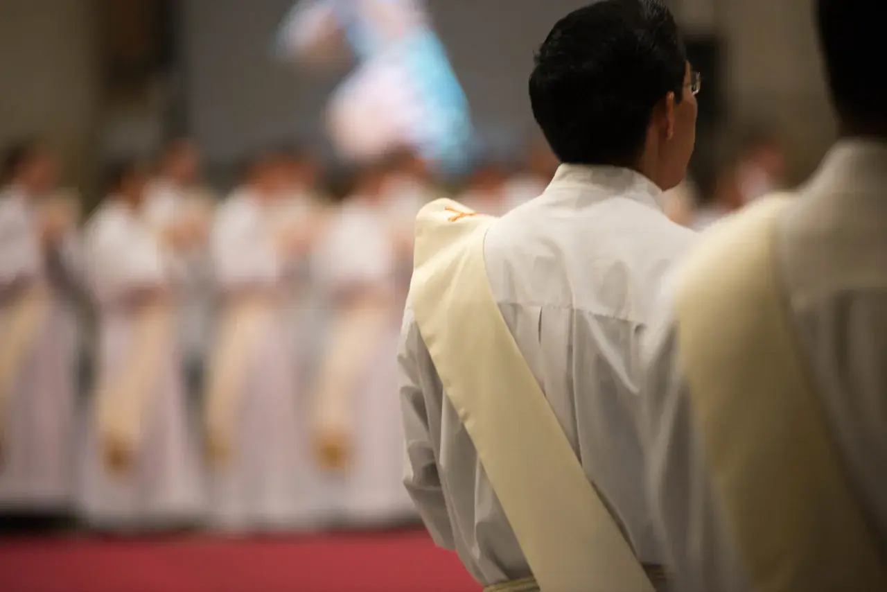 Diakon w białej szacie z żółtą stułą, przygotowuje się do uroczystości. W tle widać innych duchownych.