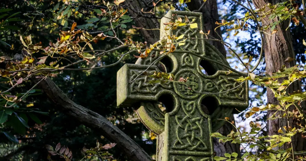 Kamienny krzyż celtycki z misternymi wzorami, otoczony jesiennymi liśćmi i gałęziami drzew.