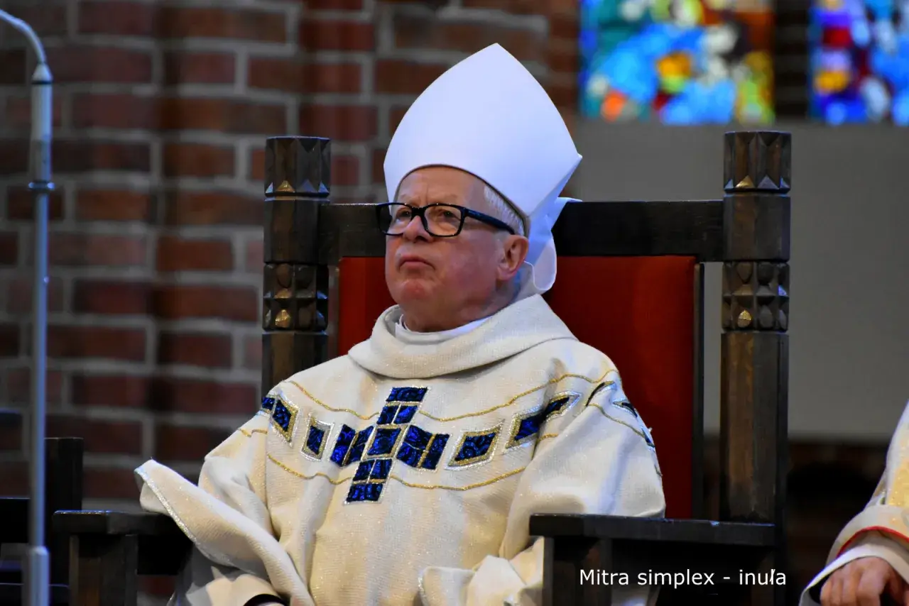 Biskup w białej mitrze i ozdobnej szacie z krzyżem, siedzi na drewnianym tronie.
