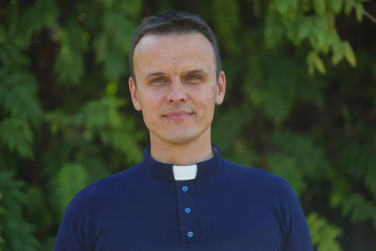 Młody ksiądz z osiedla w granatowej koszuli z kołnierzykiem, uśmiecha się na tle zieleni.