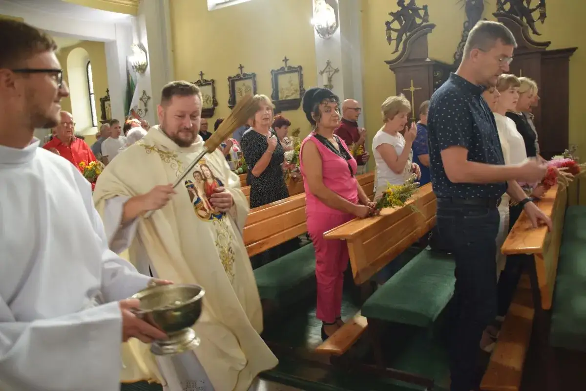 Proboszcz błogosławi wiernych w kościele, trzymając w dłoni kropidło i naczynie z wodą. Ludzie stoją w ławkach, trzymając bukiety ziół i kwiatów.