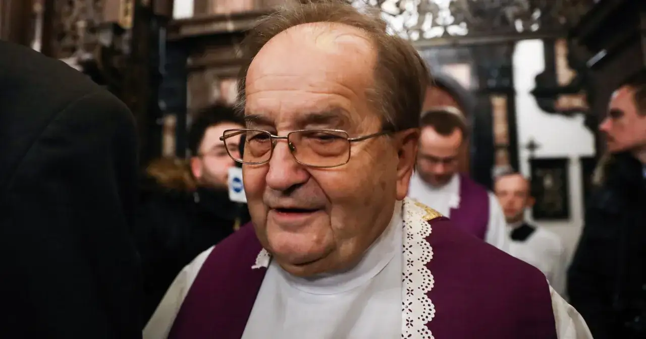 Ksiądz Rydzyk w okularach i sutannie z fioletową stułą, otoczony przez innych duchownych w kościele.