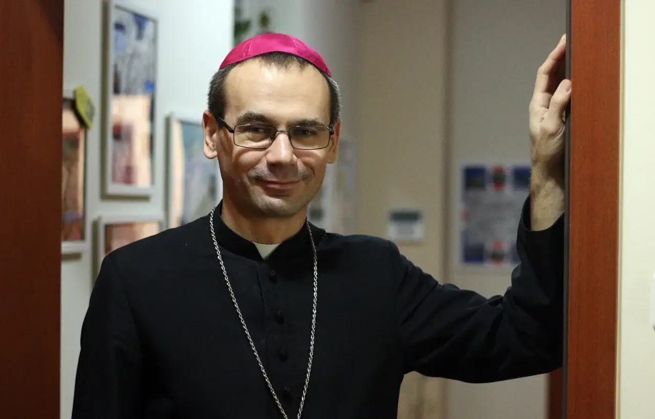 Uśmiechnięty, najmłodszy biskup w Polsce, w czarnej sutannie i z różowym zucchetto na głowie, opiera się o framugę drzwi.