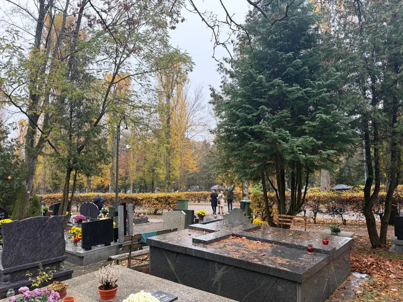 Jesienny cmentarz, grobbing co to? Widać nagrobki, drzewa i ludzi z parasolami.
