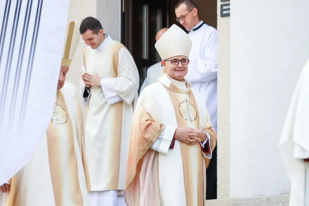 Uśmiechnięty biskup katowicki w białej mitrze i szacie z krzyżem na szyi, otoczony przez innych duchownych.