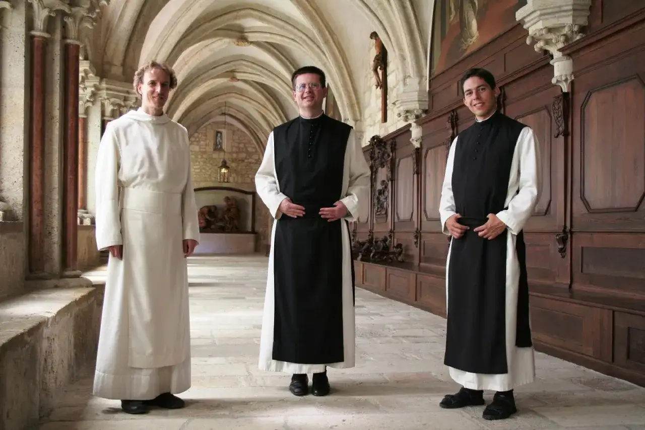 Trzech mężczyzn w habitach, jeden zakonnik w białym habicie, stoi w klasztornym korytarzu.