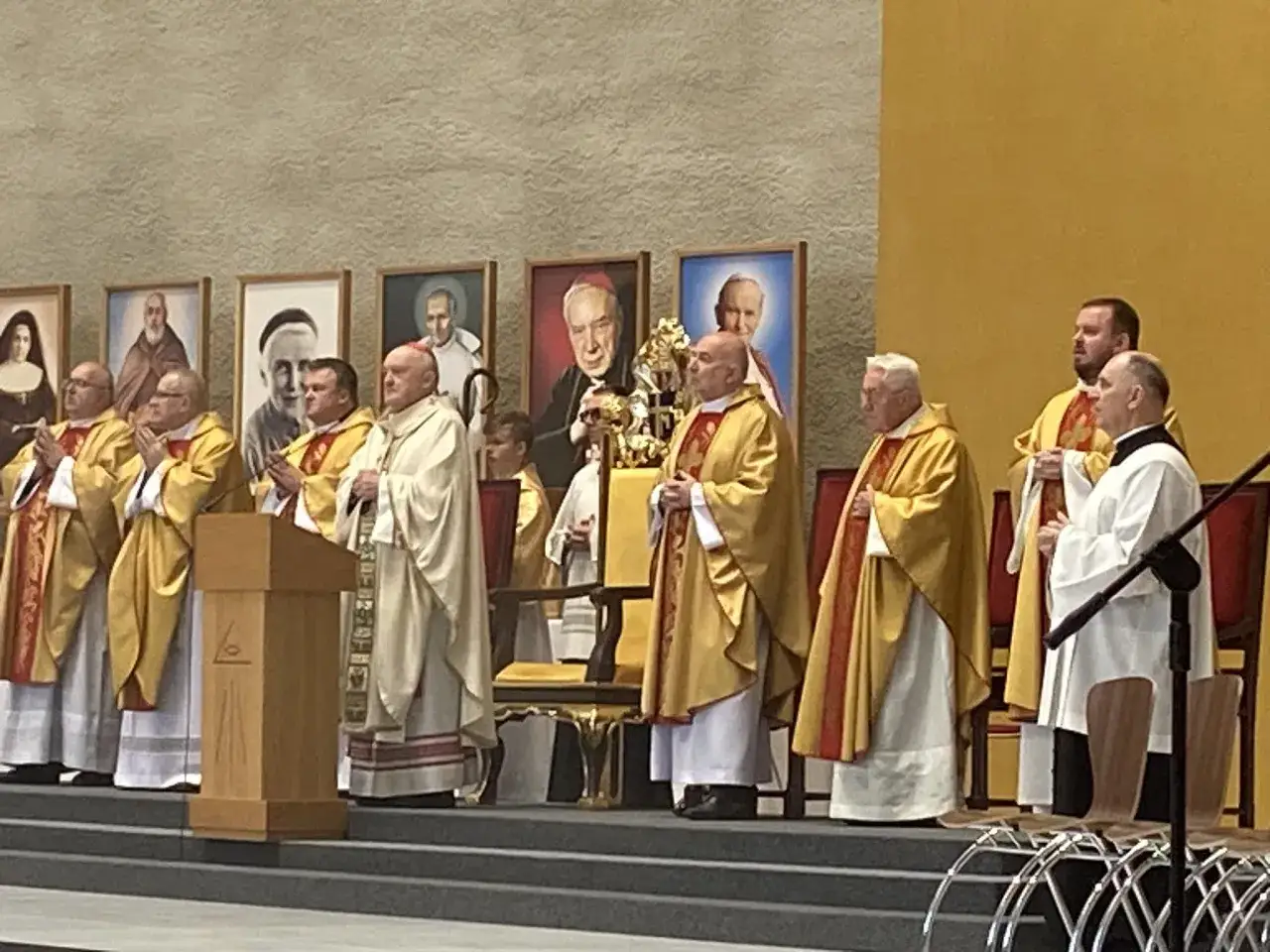 Biskup Nycz wśród duchownych w szatach liturgicznych, na tle portretów świętych i błogosławionych.