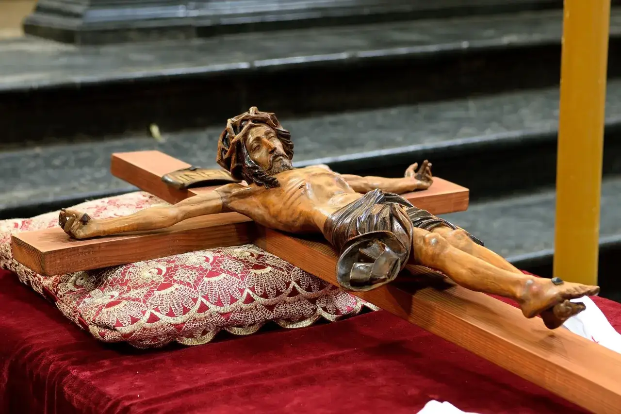 Drewniany post paschalny z Jezusem na krzyżu, ozdobiony bogato haftowaną poduszką, symbolizuje mękę i zmartwychwstanie.