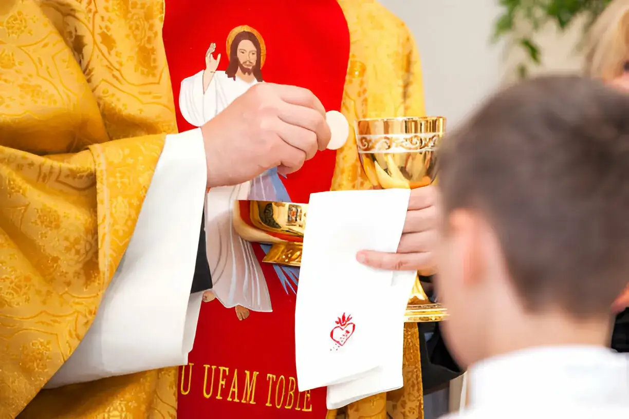 Kapłan w złotej szacie z wizerunkiem Jezusa podczas post eucharystyczny podaje hostię dziecku.