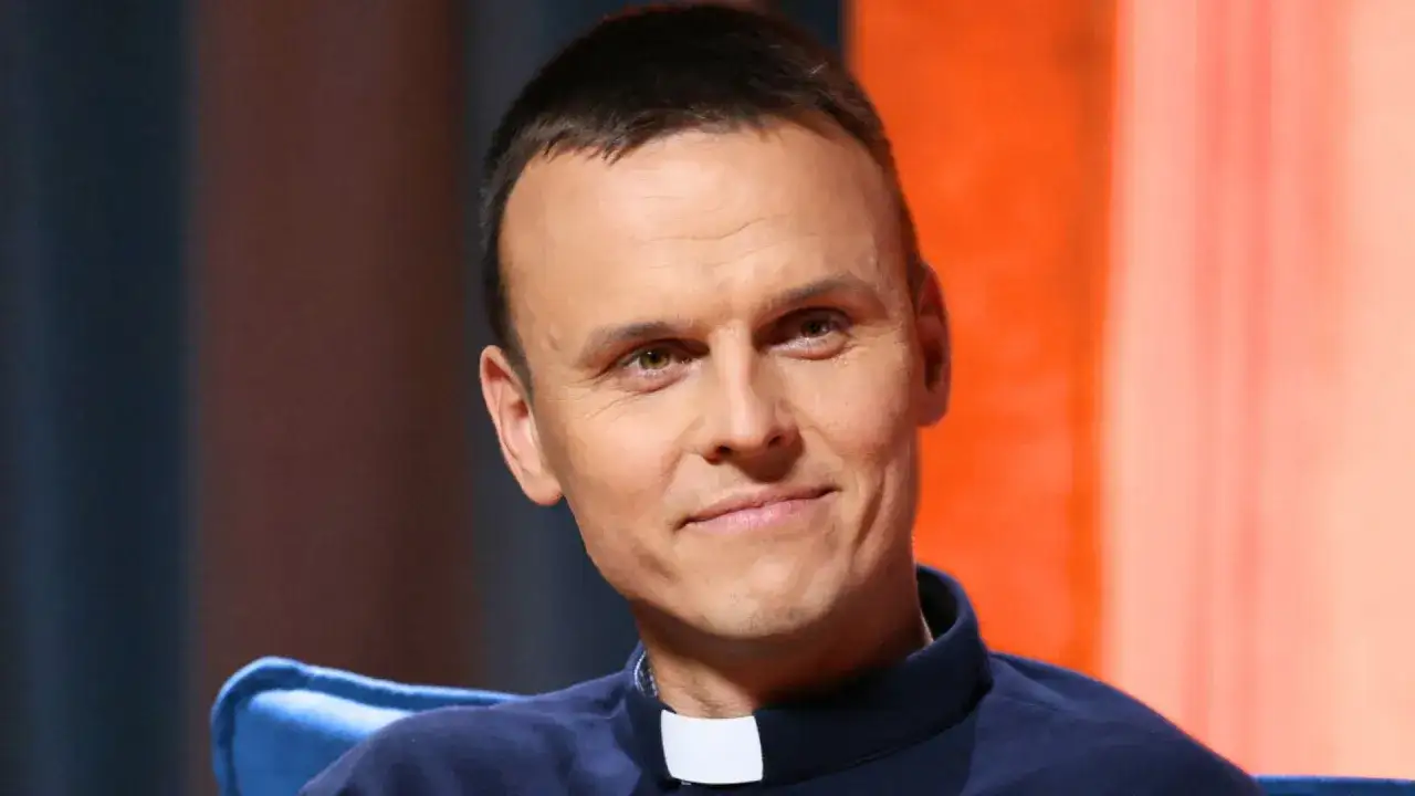 Młody ksiądz z osiedla, z uśmiechem na twarzy, w granatowej koszuli z białym kołnierzykiem.