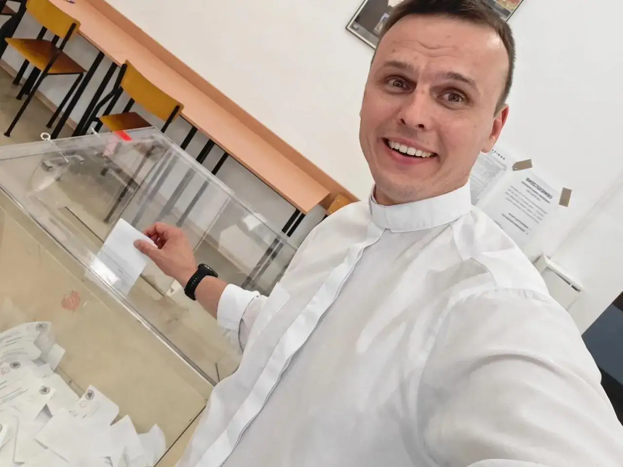 Uśmiechnięty ksiądz z osiedla wrzuca kartę do głosowania do urny.