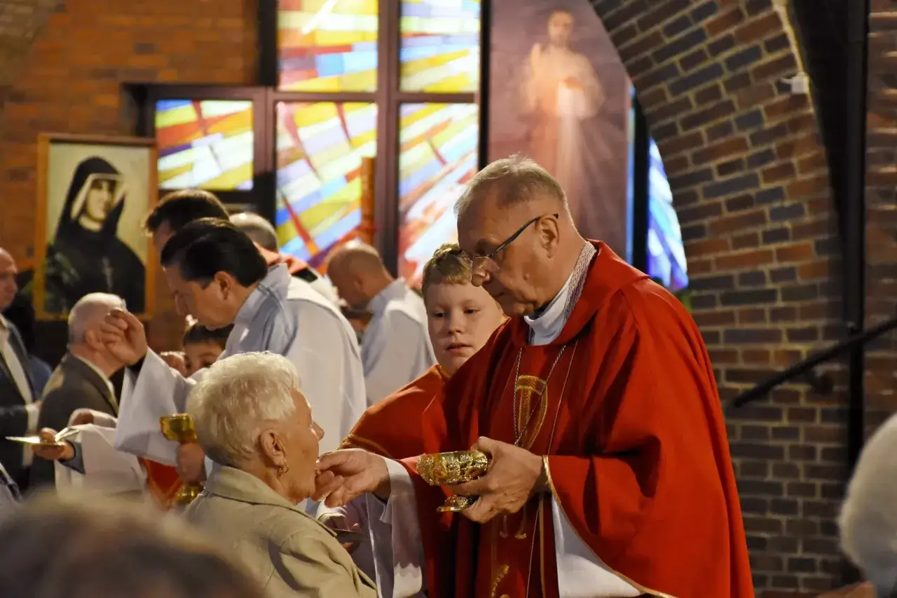 Kapłan udziela Komunii Świętej starszej pani. W tle witraże i obraz Matki Bożej. To piękny moment błogosławieństwa.