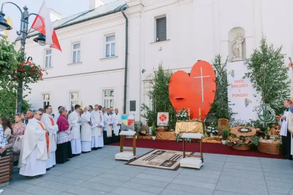 Uroczysta procesja Bożego Ciała przed kościołem. Wierni w białych szatach, wielkie serce z krzyżem i transparent "Król Miłości i Pokoju".